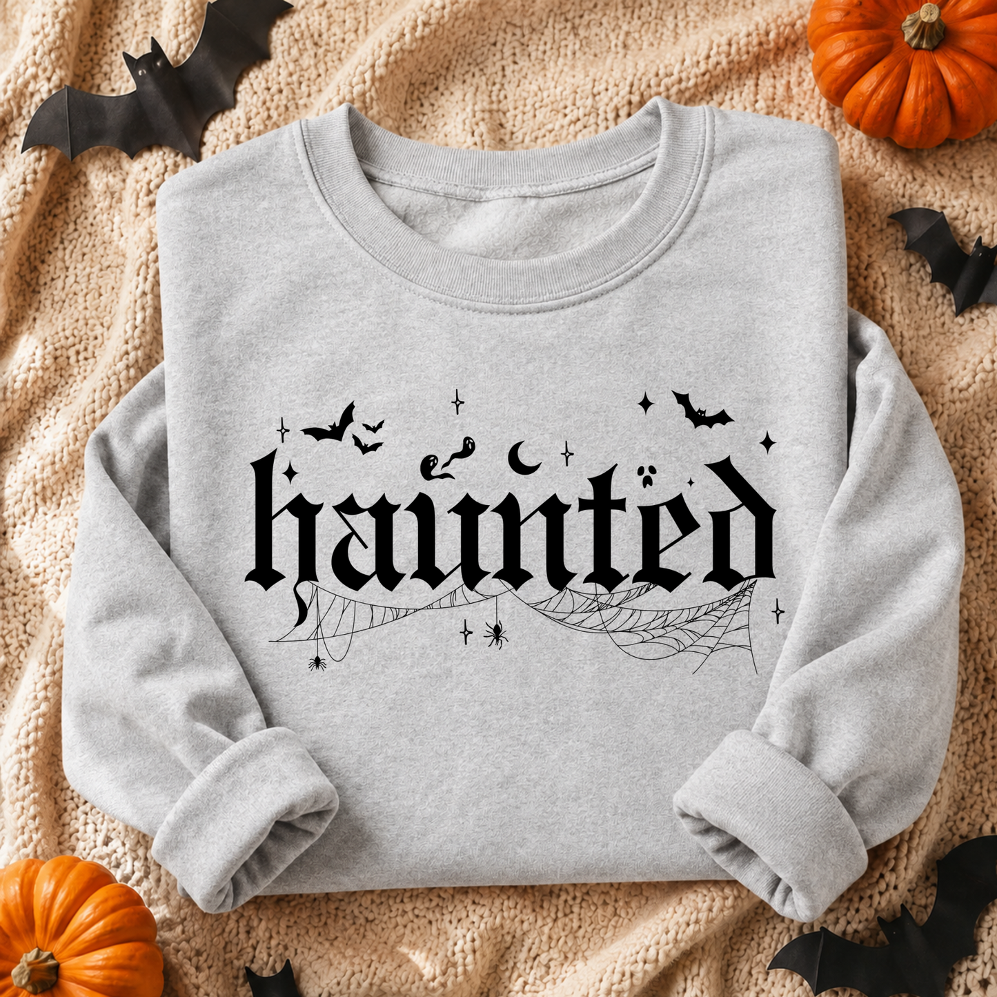 A light gray crewneck sweatshirt with a black Gothic-style 'haunted' graphic, styled in a flat lay on a beige knit blanket with Halloween decorations including pumpkins, bats, and spiderwebs.