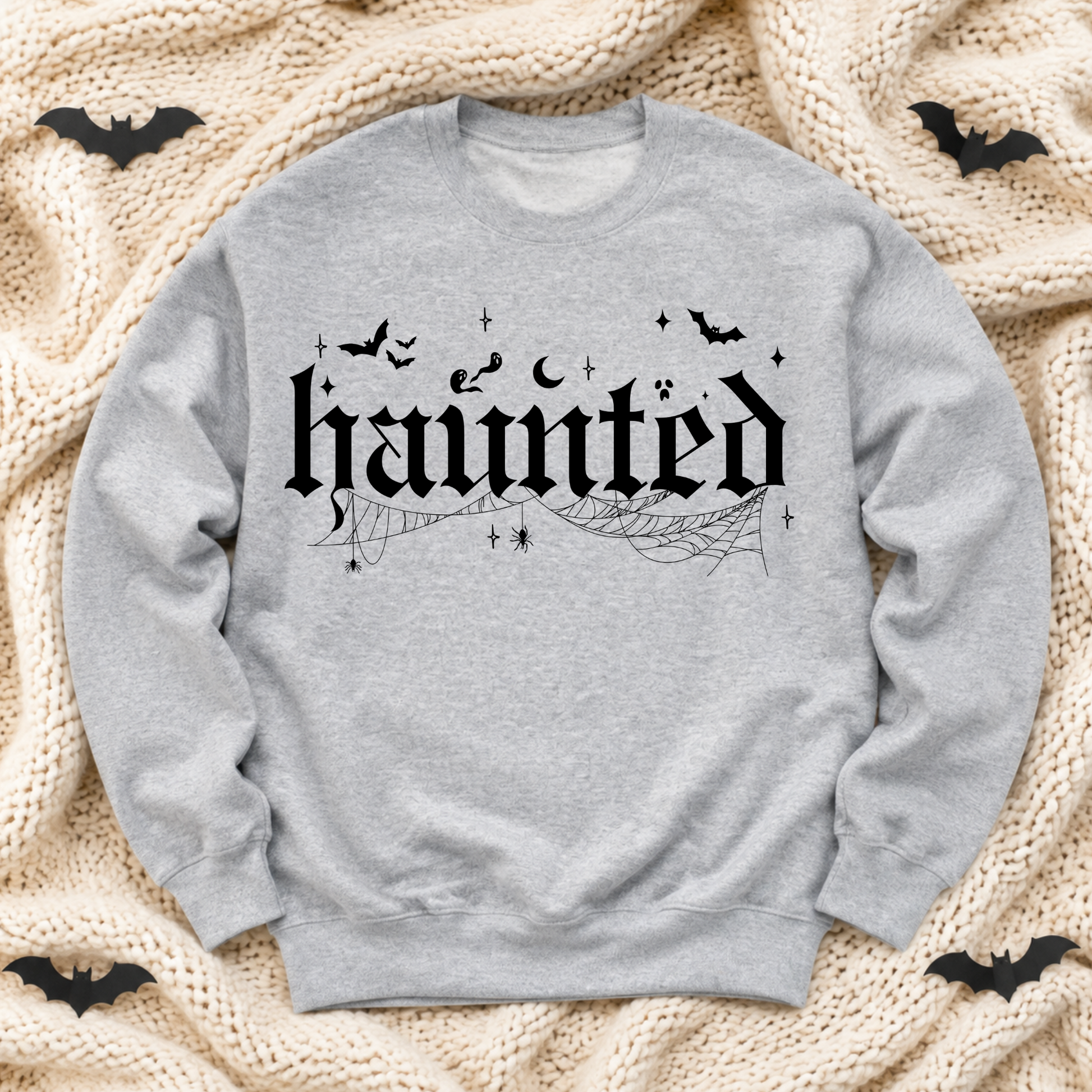 A light ash gray crewneck sweatshirt with a black Gothic-style 'haunted' graphic, spiderwebs, moons, and bats, laid flat on a beige knit blanket with small black bat cutouts in the corners.