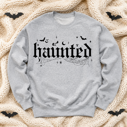 A light ash gray crewneck sweatshirt with a black Gothic-style 'haunted' graphic, spiderwebs, moons, and bats, laid flat on a beige knit blanket with small black bat cutouts in the corners.