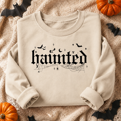 A beige sand crewneck sweatshirt with a black gothic-style 'haunted' graphic, spiderwebs, bats, and stars, presented in a flat-lay Halloween setting with pumpkins, black bats, and a knitted blanket.