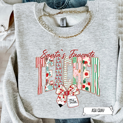 Ash gray Santa’s Favorite Teacher sweatshirt with festive striped lettering and holiday bow, styled in a flat lay