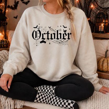 A woman wearing a cream-colored crewneck sweatshirt with a black gothic 'October' graphic featuring bats, spiders, and cobwebs, sitting in a cozy, decorated Halloween-themed room.