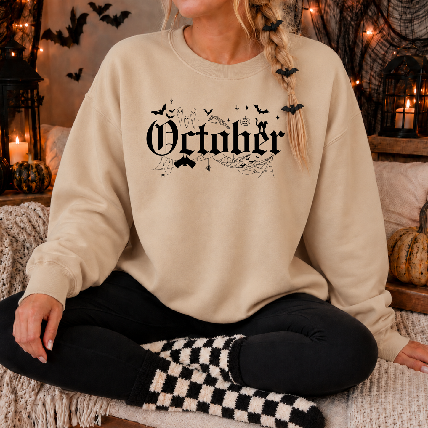 A woman wearing a beige crewneck sweatshirt with a black gothic 'October' graphic featuring bats, a witch, and a cauldron, sitting in a cozy, Halloween-themed room with candles and pumpkins.