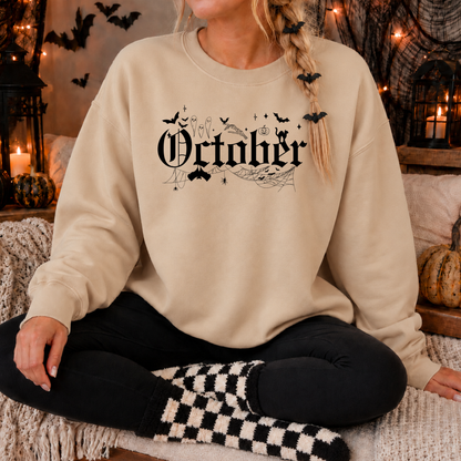 A woman wearing a beige crewneck sweatshirt with a black gothic 'October' graphic featuring bats, a witch, and a cauldron, sitting in a cozy, Halloween-themed room with candles and pumpkins.