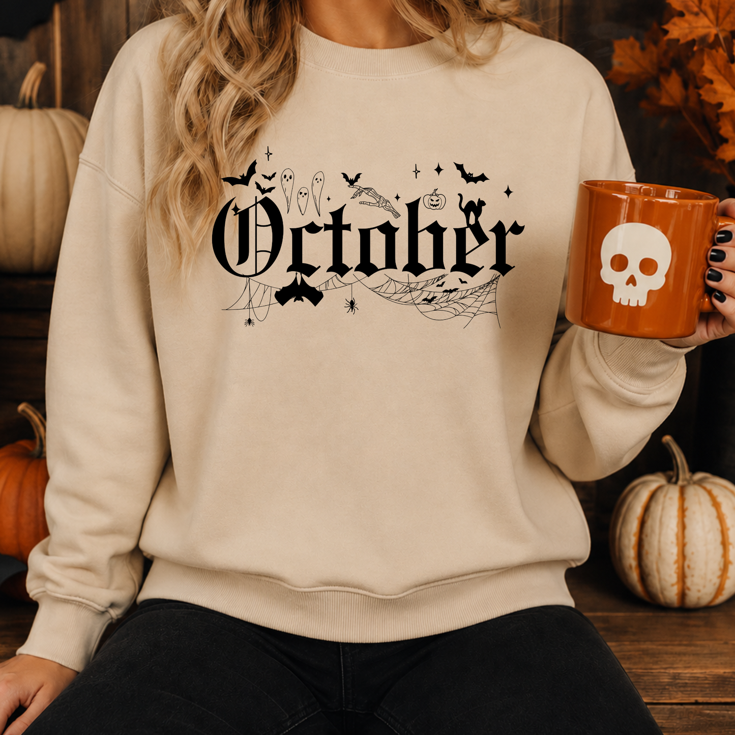 A woman wearing a cream-colored crewneck sweatshirt with a black 'October' gothic graphic and Halloween motifs, holding an orange mug with a white skull, set against a rustic autumn background with pumpkins.