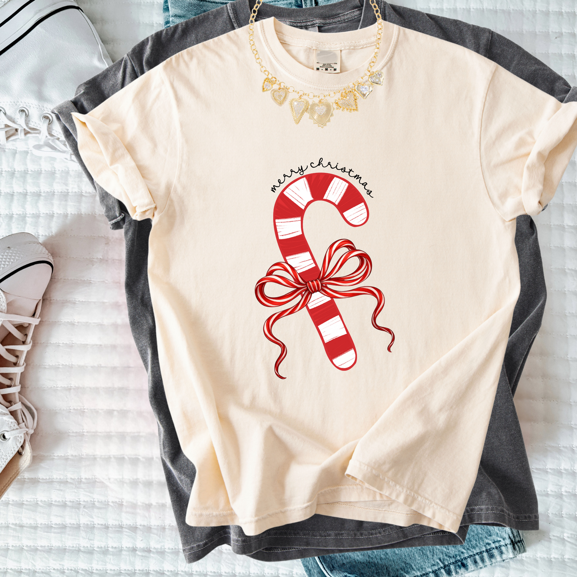 Cream-colored t-shirt with a red candy cane and ribbon Merry Christmas design laid flat with layered clothing.