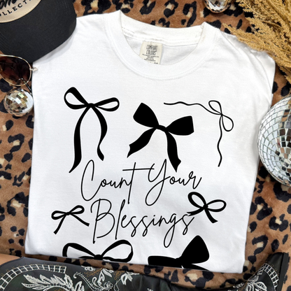 White t-shirt with black bows and 'Count Your Blessings' text on a leopard print surface.