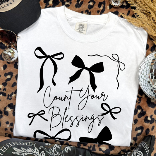 White t-shirt with black bows and 'Count Your Blessings' text on a leopard print surface.