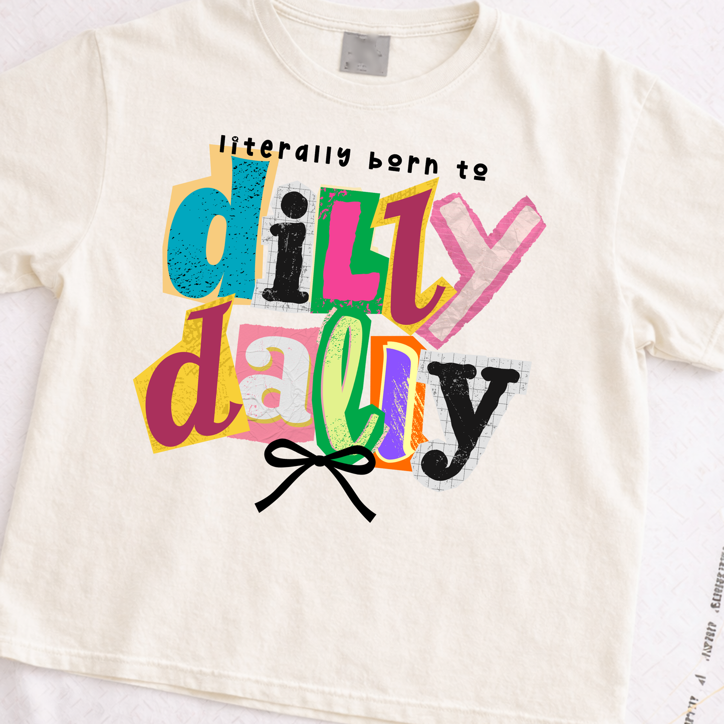 ivory t-shirt with colorful text 'literally born to dilly dally' and a bow graphic on a white background.