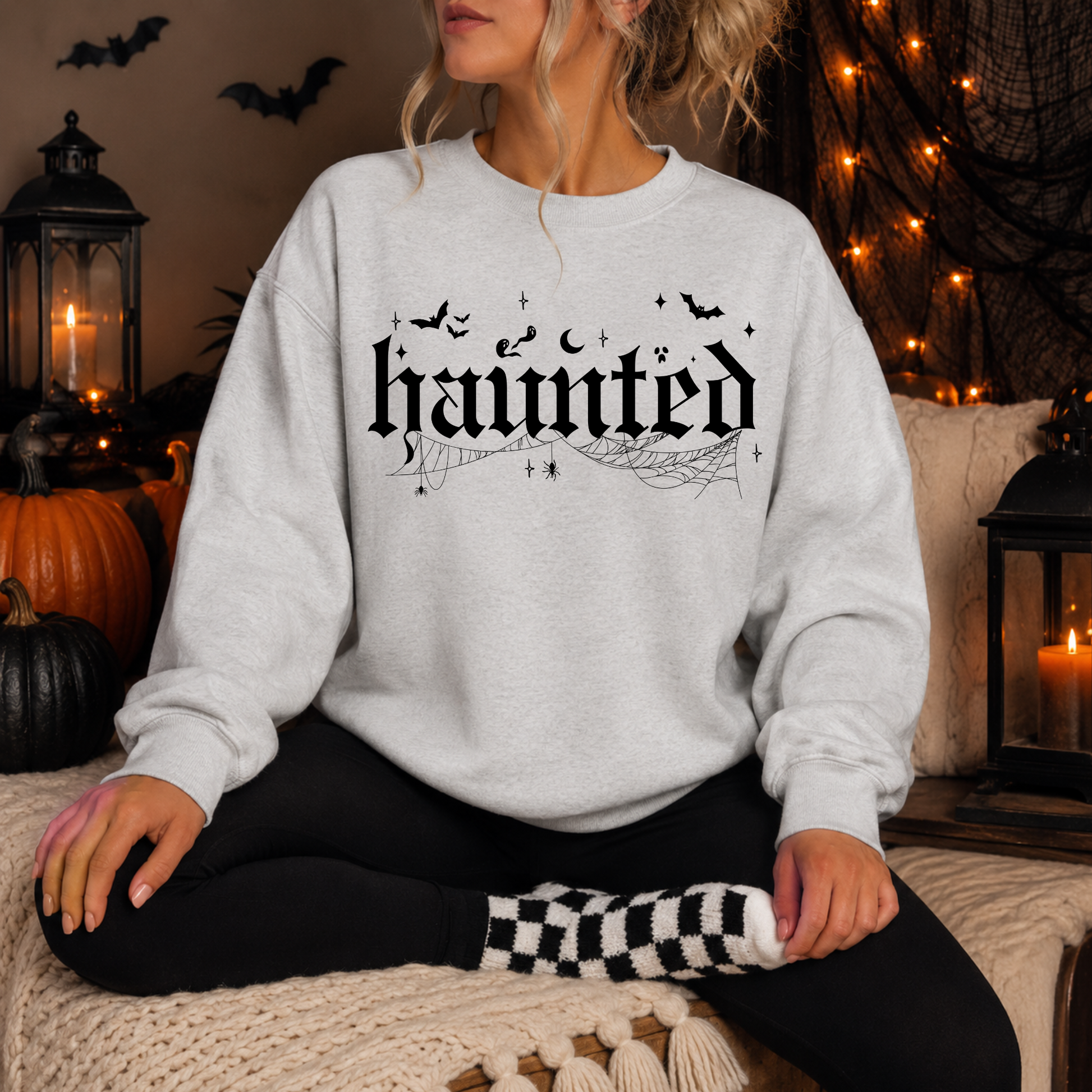 A woman wearing a light gray sweatshirt with a black gothic-style 'haunted' graphic sits cross-legged in a cozy, Halloween-themed room decorated with pumpkins, string lights, and lanterns.