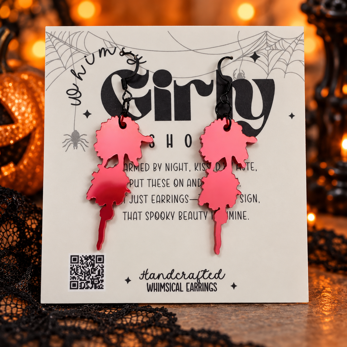 Handcrafted whimsical earrings shaped like two overlapping pink and red witch hats on a white display card, set against a dark, spooky background with black lace and warm orange bokeh lights.