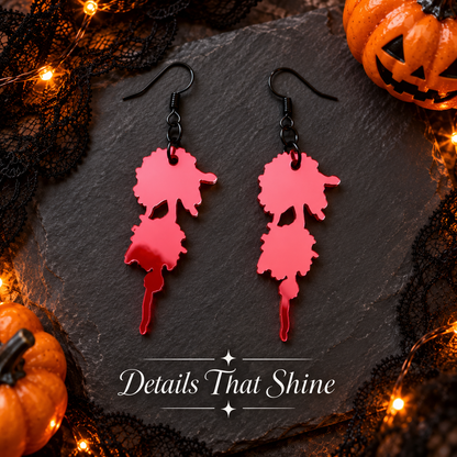 A pair of dangling earrings with bright pink and red dripping paint shapes on black hooks, displayed on a dark slate board with Halloween props including black lace, string lights, and small pumpkins. The text 'Details That Shine' is visible at the bottom.