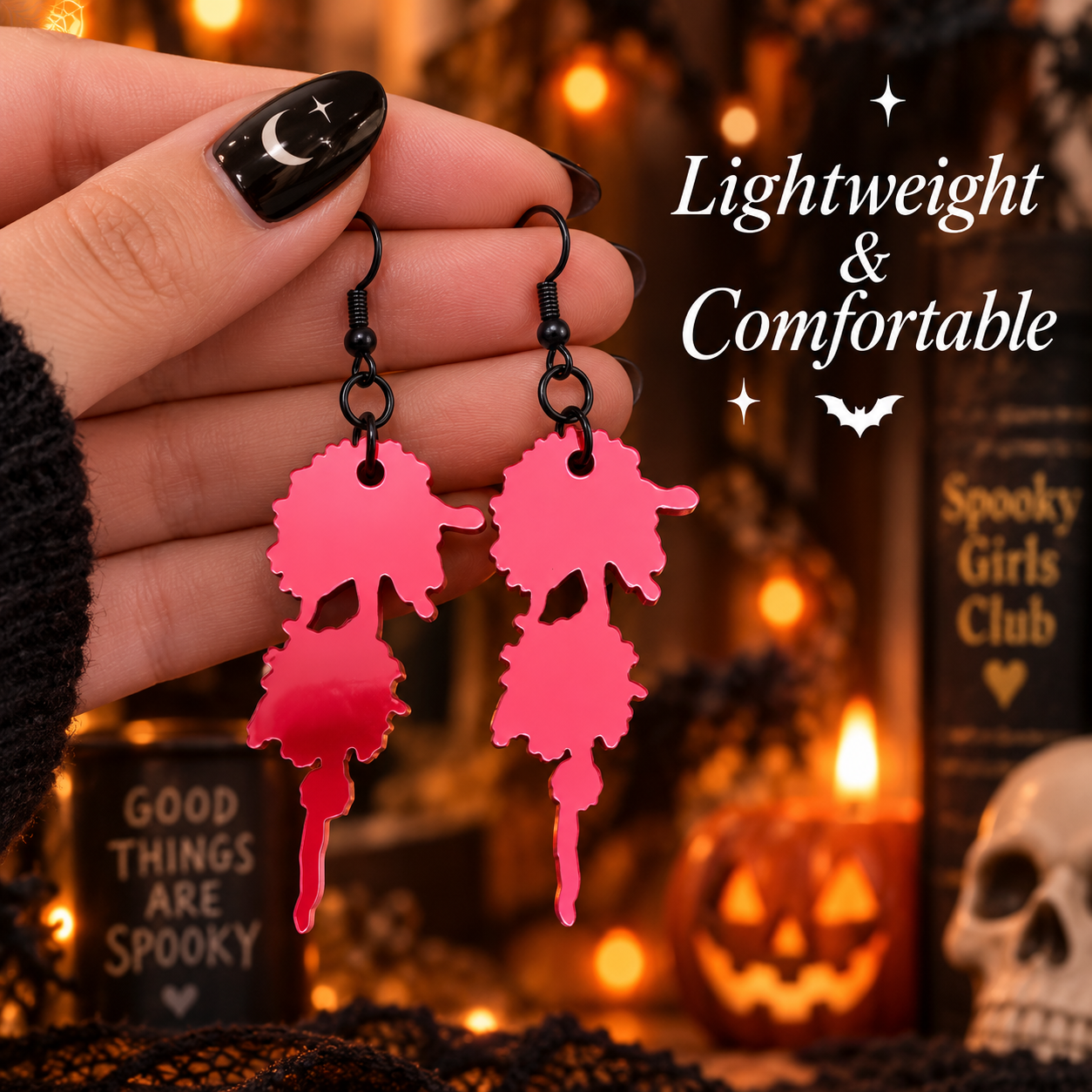 A hand with black nail art holds a pair of dangling earrings shaped like dripping blood splatters in a bright pink metallic finish. The background is a cozy, out-of-focus Halloween scene with warm lights, a skull, and a book reading 'Spooky Girls Club'.