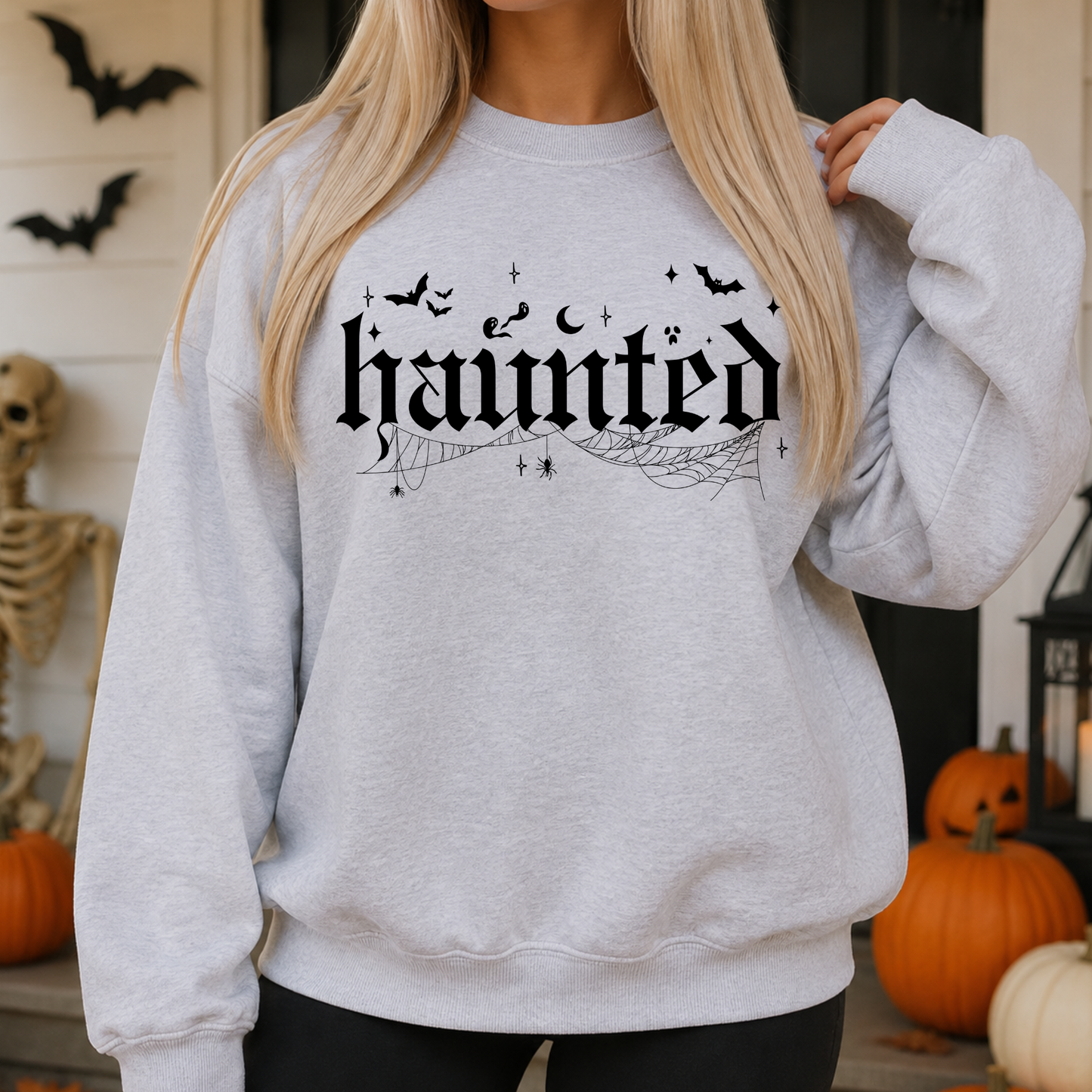 A woman wearing a light gray crewneck sweatshirt with a black gothic-style 'haunted' graphic and spiderwebs, posed in a Halloween-themed setting with pumpkins and bats.