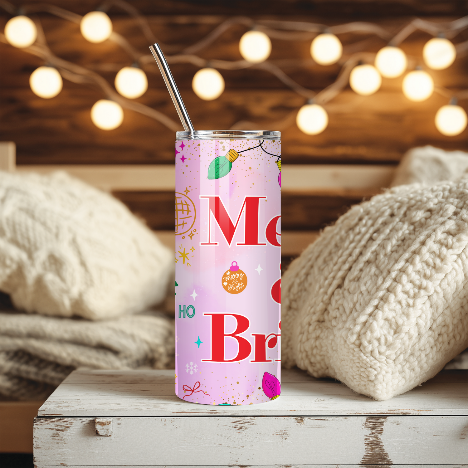 Pink “Merry & Bright” Christmas tumbler on a cozy holiday background with warm string lights and knitted blankets. Stainless steel 20 oz cup with festive pastel ornaments.