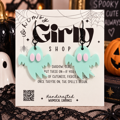 Whimsical ghost-shaped drop earrings in mint green with pink eyes, displayed on a branded 'We Numsy Girly Shop' card against a Halloween-themed background.