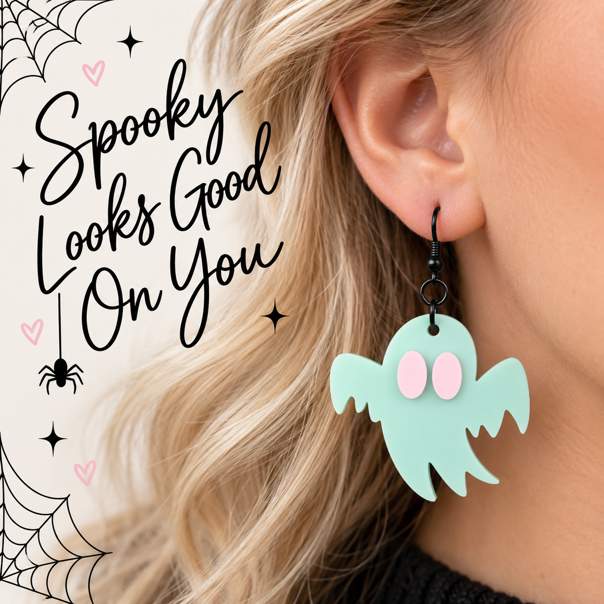 A close-up of a person's ear wearing a mint green ghost-shaped dangle earring with pink eyes and black hooks. The person has blonde hair, and the background is a light cream color with Halloween-themed graphic text reading 'Spooky Looks Good On You' and spider/web illustrations.
