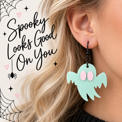 A close-up of a person's ear wearing a mint green ghost-shaped dangle earring with pink eyes and black hooks. The person has blonde hair, and the background is a light cream color with Halloween-themed graphic text reading 'Spooky Looks Good On You' and spider/web illustrations.