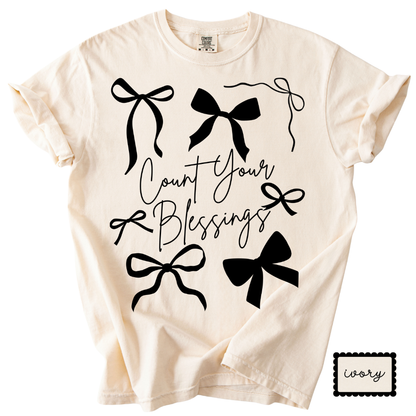 Beige t-shirt with black bows and 'Count Your Blessings' text on a white background