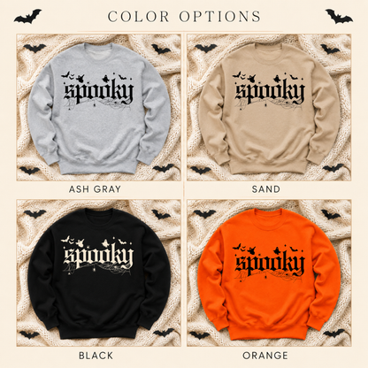Four color options for a crewneck sweatshirt with a gothic 'spooky' graphic: Ash Gray, Sand, Black, and Orange, each shown on a textured beige background with black bat silhouettes.