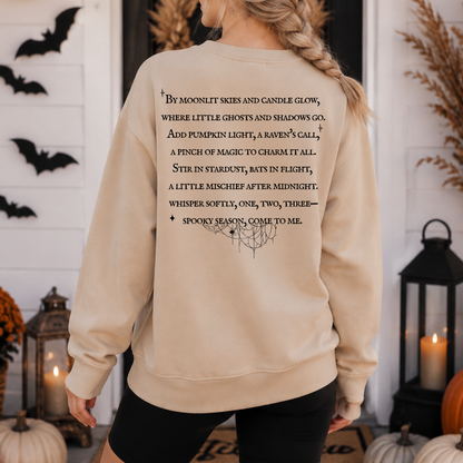 A woman wearing a beige crewneck sweatshirt with a black printed spooky poem on the back, shown from behind in a decorated Halloween-themed setting with pumpkins, lanterns, and bats.