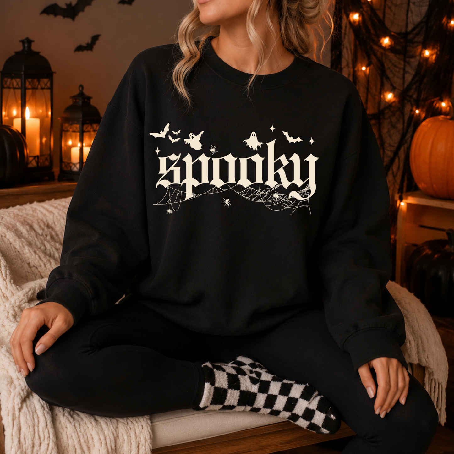 A woman sits cross-legged on a couch wearing a black crewneck sweatshirt with a white 'spooky' graphic featuring bats, ghosts, and cobwebs. The background is a cozy, warmly lit Halloween setting with candles, lanterns, and pumpkins.