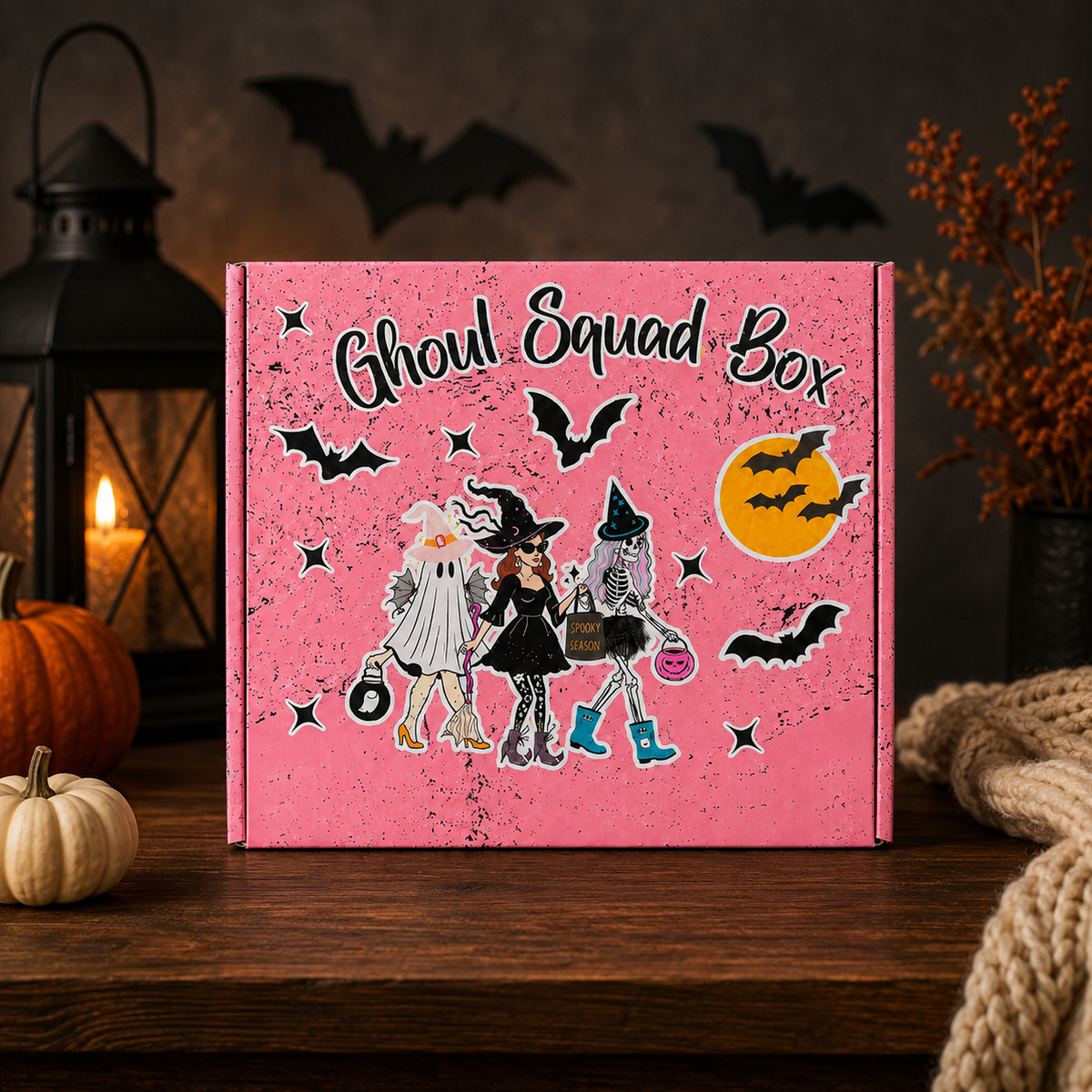 A pink 'Ghoul Squad Box' product box decorated with Halloween illustrations, displayed on a rustic wooden table with seasonal props like a lantern, pumpkins, and a rope.
