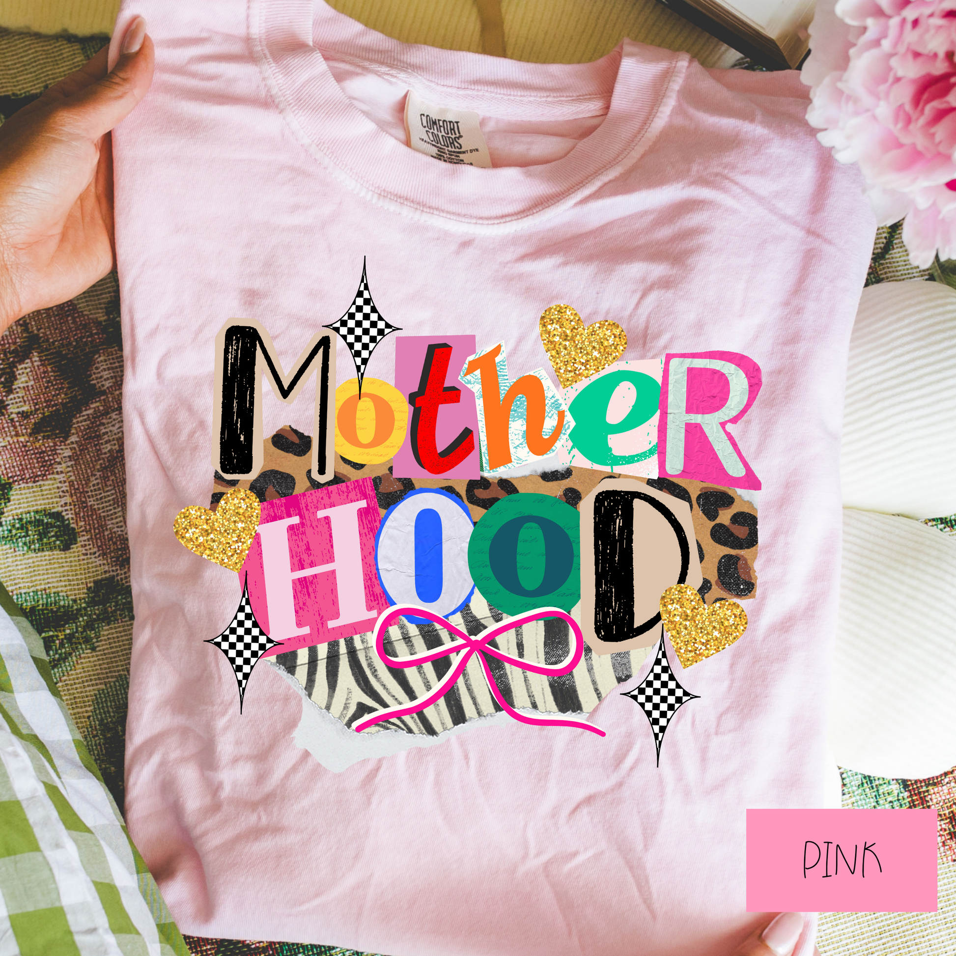 Pink t-shirt with colorful 'Mother Hood' text and decorative elements, held by a hand.