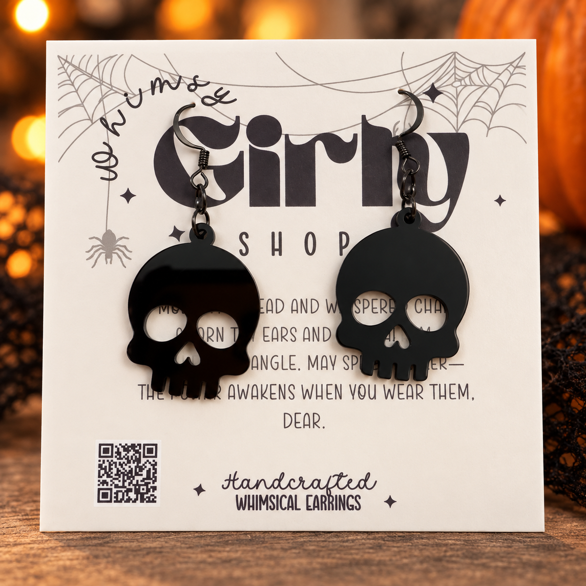 A pair of black skull-shaped dangling earrings displayed on a white printed card that reads 'Whimsy Girly Shop' and 'Handcrafted Whimsical Earrings.' The card is photographed on a wooden surface with warm blurred lights in the background.