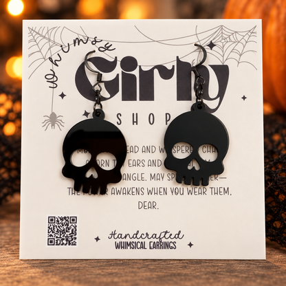 A pair of black skull-shaped dangling earrings displayed on a white printed card that reads 'Whimsy Girly Shop' and 'Handcrafted Whimsical Earrings.' The card is photographed on a wooden surface with warm blurred lights in the background.
