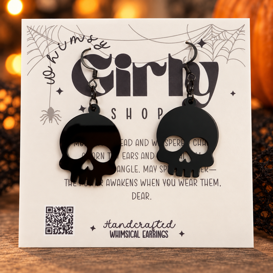 A pair of black skull-shaped dangling earrings displayed on a white printed card that reads 'Whimsy Girly Shop' and 'Handcrafted Whimsical Earrings.' The card is photographed on a wooden surface with warm blurred lights in the background.
