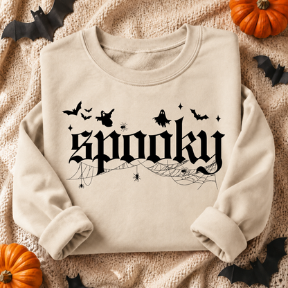 A beige crewneck sweatshirt with a black Gothic-style 'spooky' graphic, featuring silhouettes of bats, ghosts, and cobwebs, laid flat on a textured cream blanket with Halloween props like pumpkins and black bat decorations.